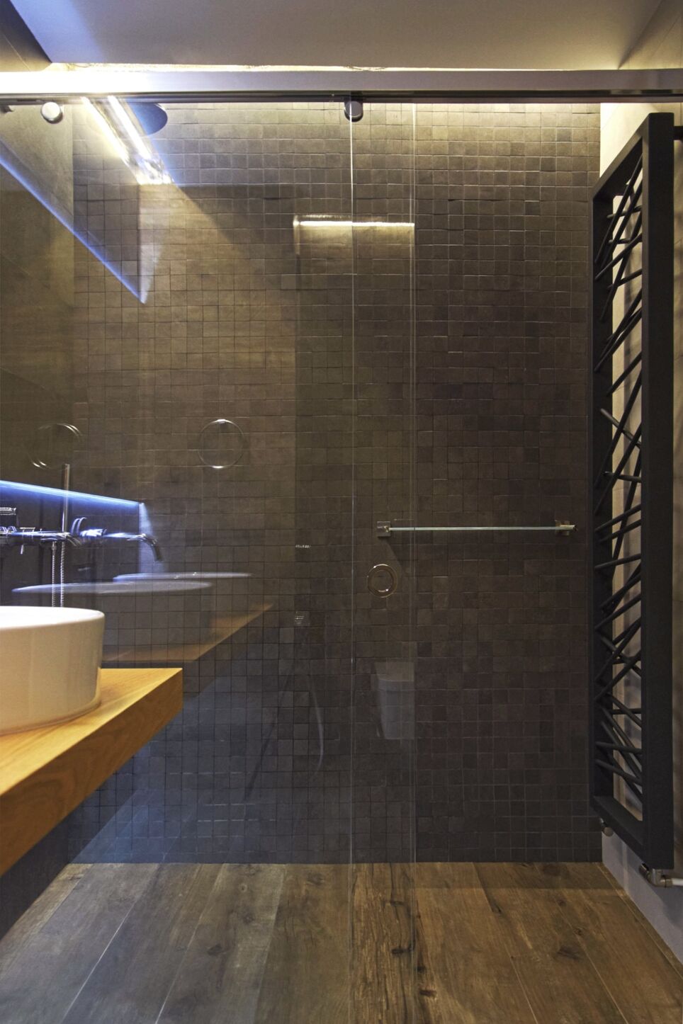 Die Glasdusche mit dunklem Mosaik und Heizleiste neben dem Eichenbrett.