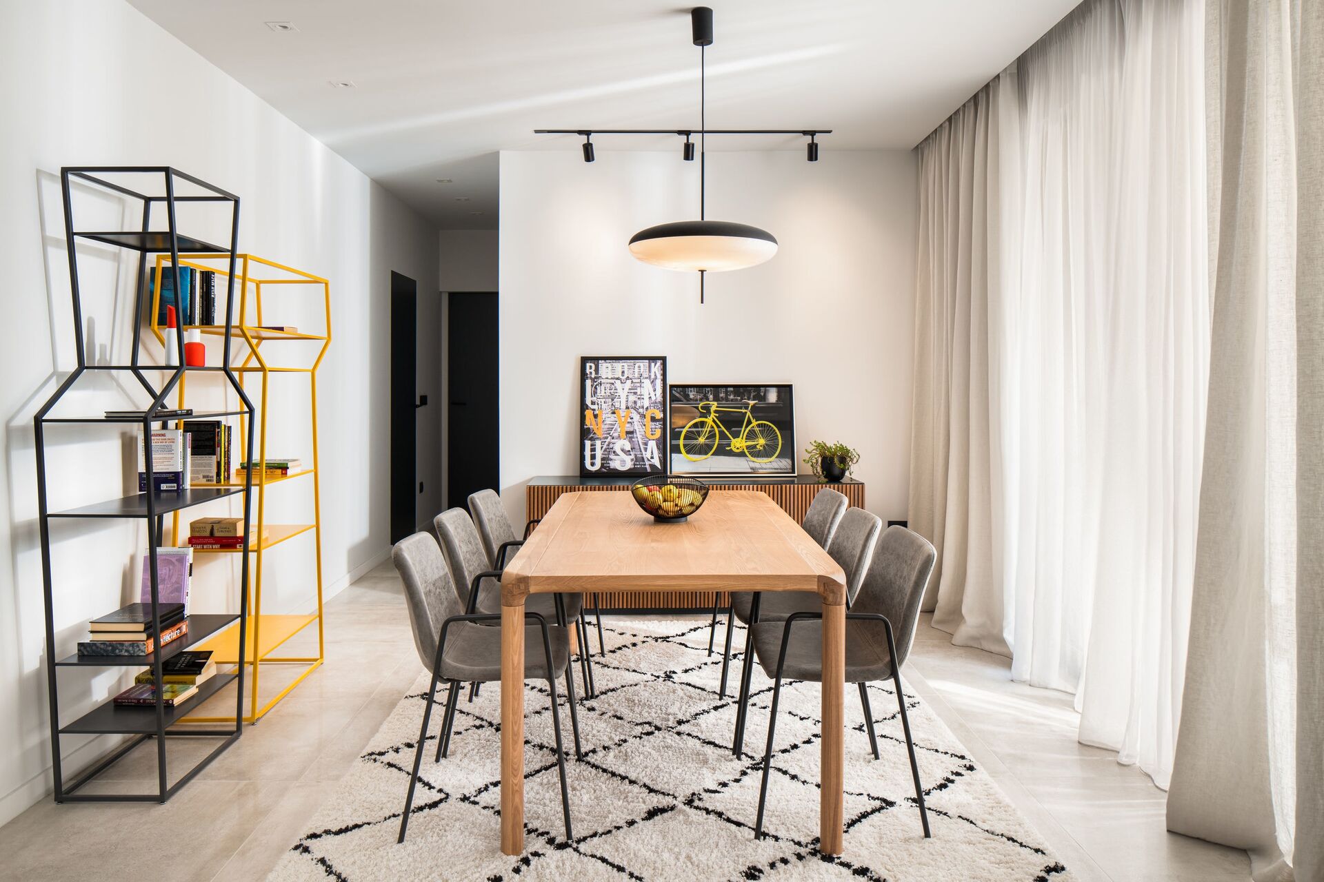 Dining room with a timber table, grey chairs and a yellow metal bookshelf by the window.