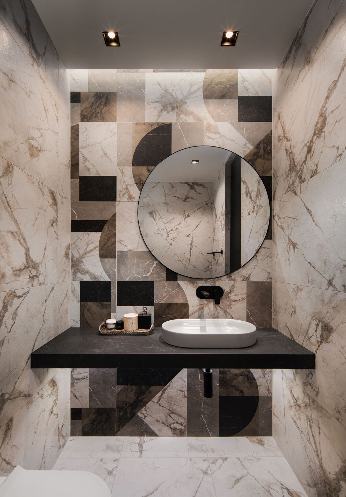 Ein WC mit geometrischen Marmorfliesen, rundem Spiegel und Waschbecken auf schwarzem Bord.